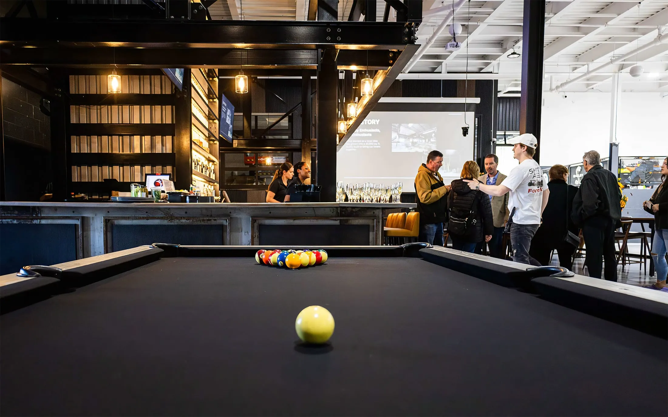 A close-up of a pool table with people chatting in the background around a bar