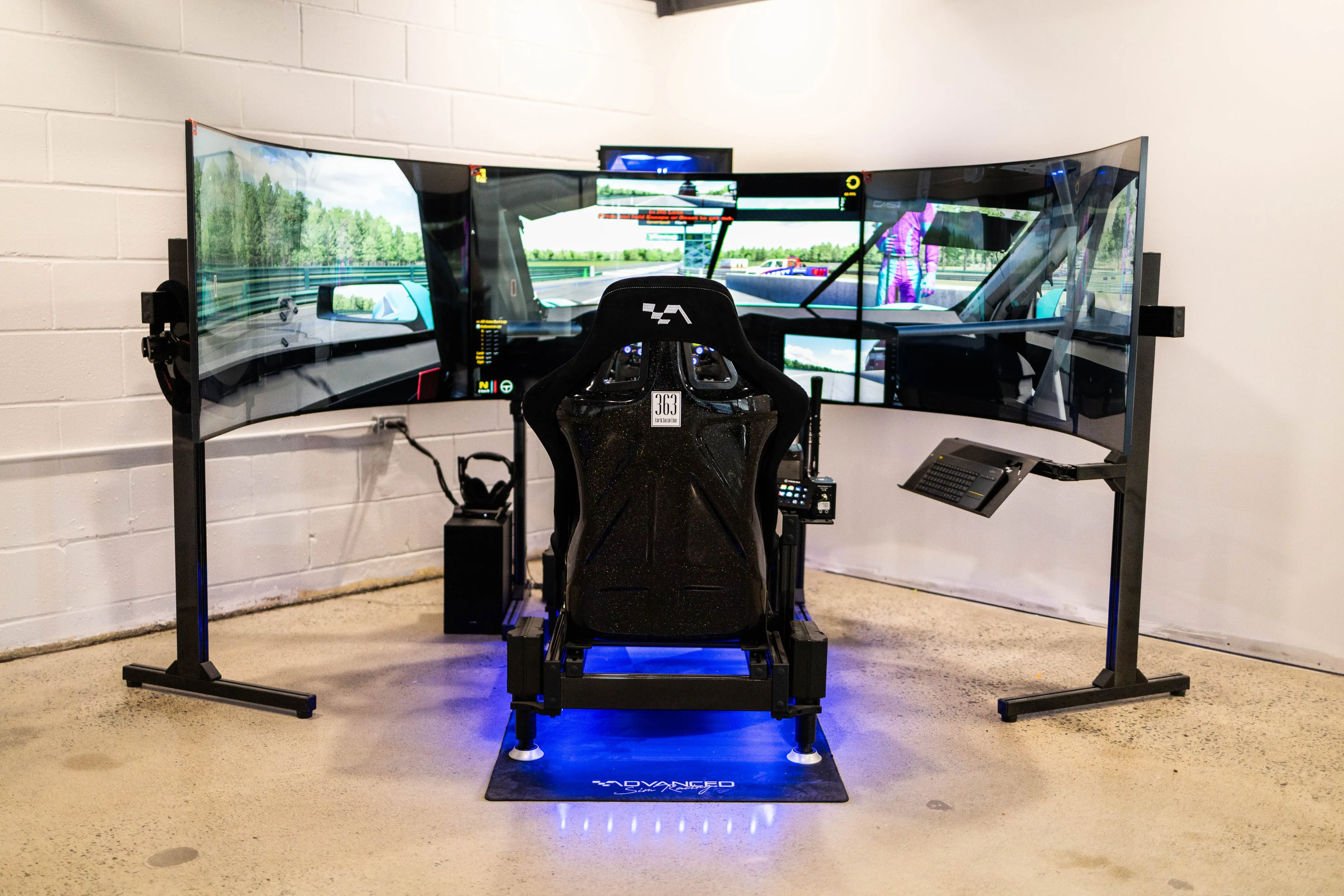 An empty racing seat with a steering wheel and pedals in front of an array of screens with a racing simulator on the main screen